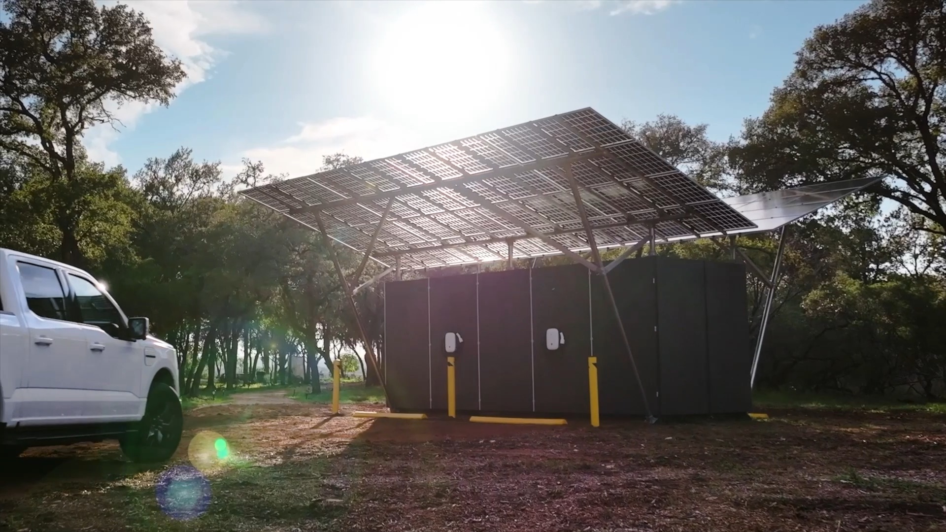 A REV unit under its solar canopy among oak trees, an electric pickup beside it